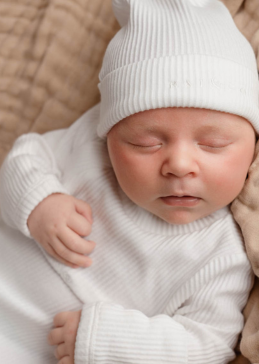 Newborn baby peacefully sleeping in a white ribbed outfit and matching beanie, resting on a soft beige blanket.