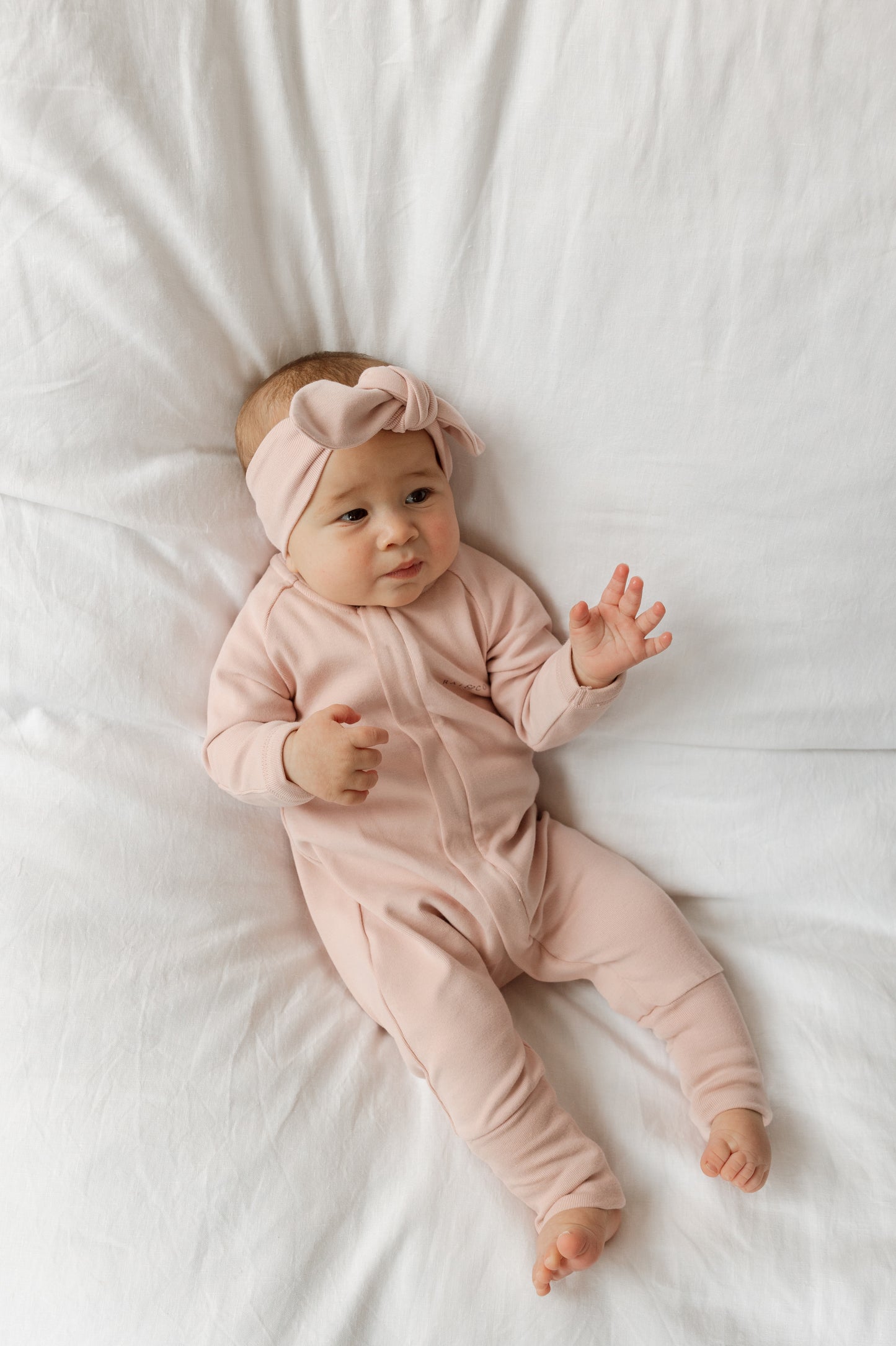A baby lying on a white surface wearing a Apple Headbands in Baby Pink  