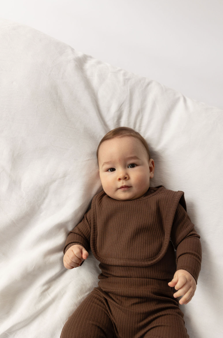 A baby lying on a white surface wearing Basic Bib Chocolate 