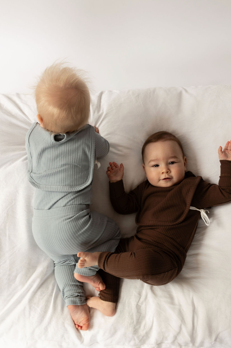 two babies wearing a Basic Bib in Chocolate and Dusty Blue on a white bed