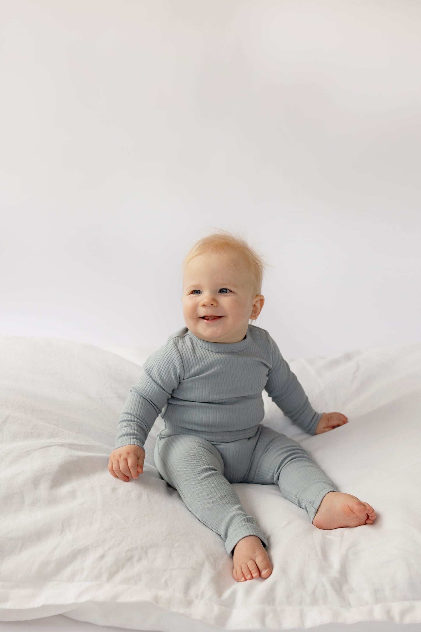 Baby wearing a Basic Leggings in Dusty Blue, sitting on a white bed 
