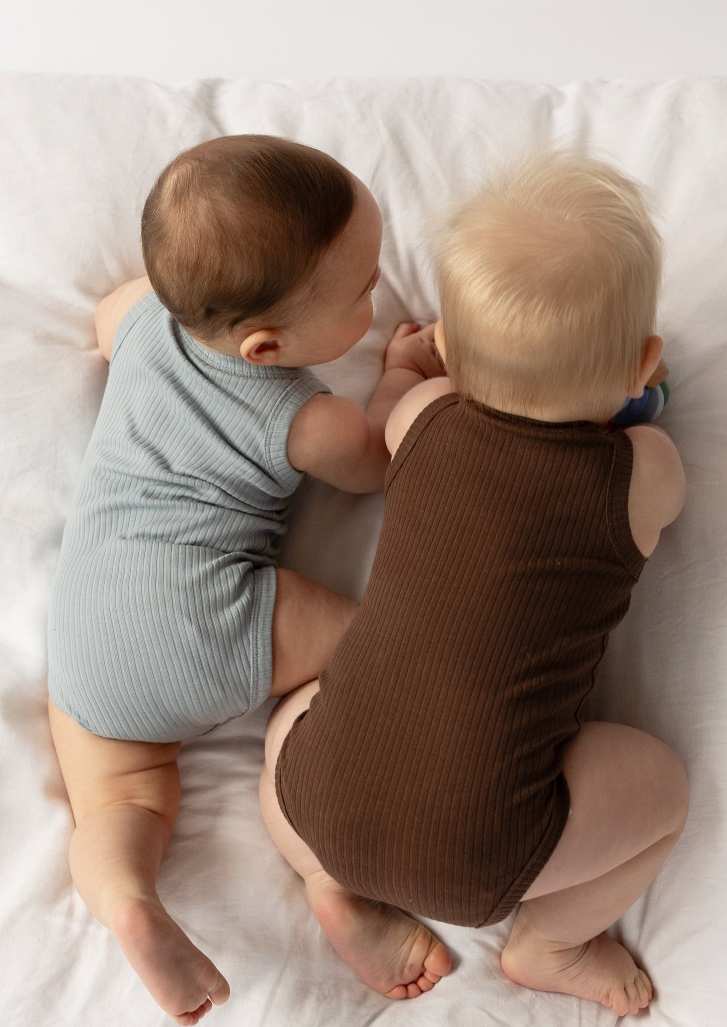 Two infants wearing a Basic Singlet Bodysuit in Dusty Blue and Chocolate, lying on their sides next to each other