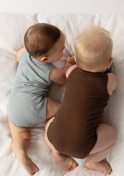 Two infants wearing a Basic Singlet Bodysuit in Dusty Blue and Chocolate, lying on their sides next to each other