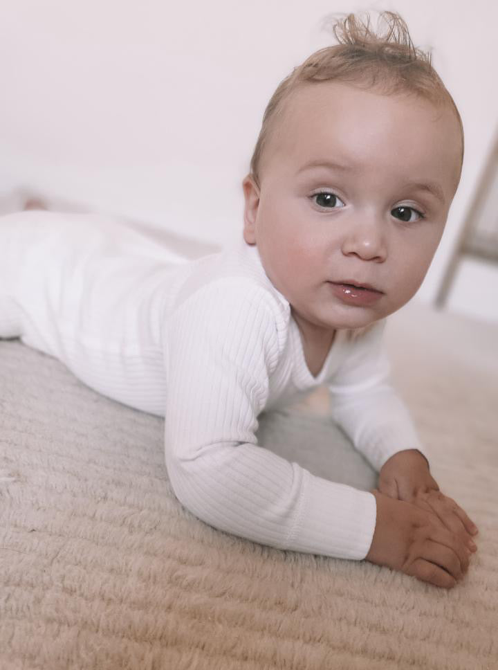 A baby wearing the Basics Long Sleeve Bodysuit in White, lying on the floor.