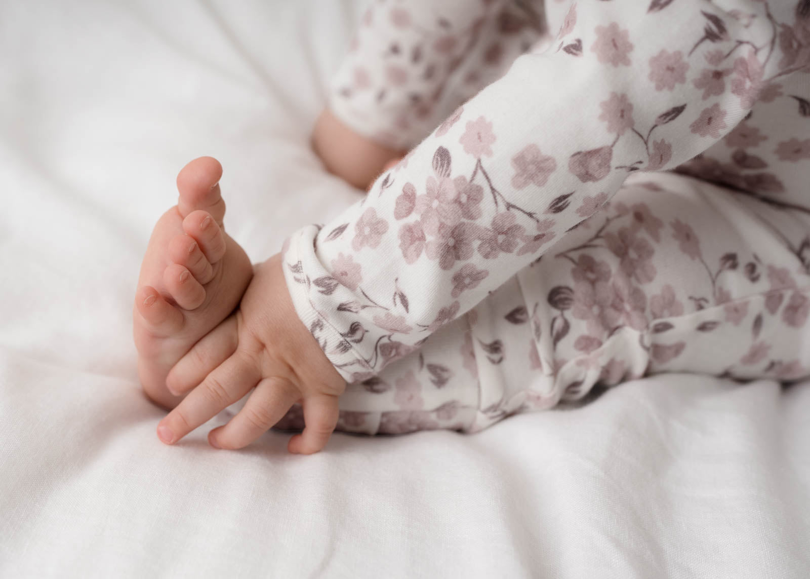 Close-up of a child's hands and feet wearing Bloom Floral Onesie Pink on a white bed.