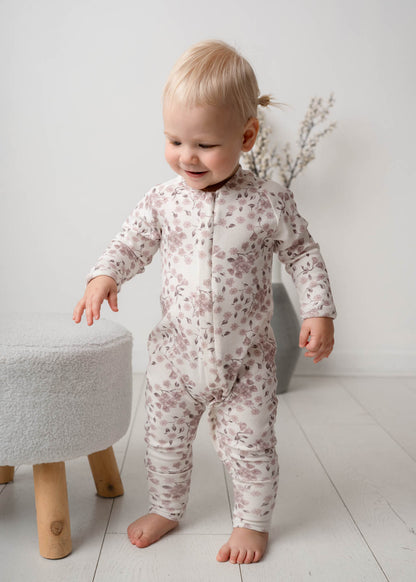 Baby in a Bloom Floral Onesie Pink standing on a light wooden floor with a white wall background.