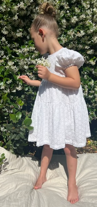A young girl wearing a Eliza Embroidered Lace Dress in White and a tie-up back standing in front of a bush.