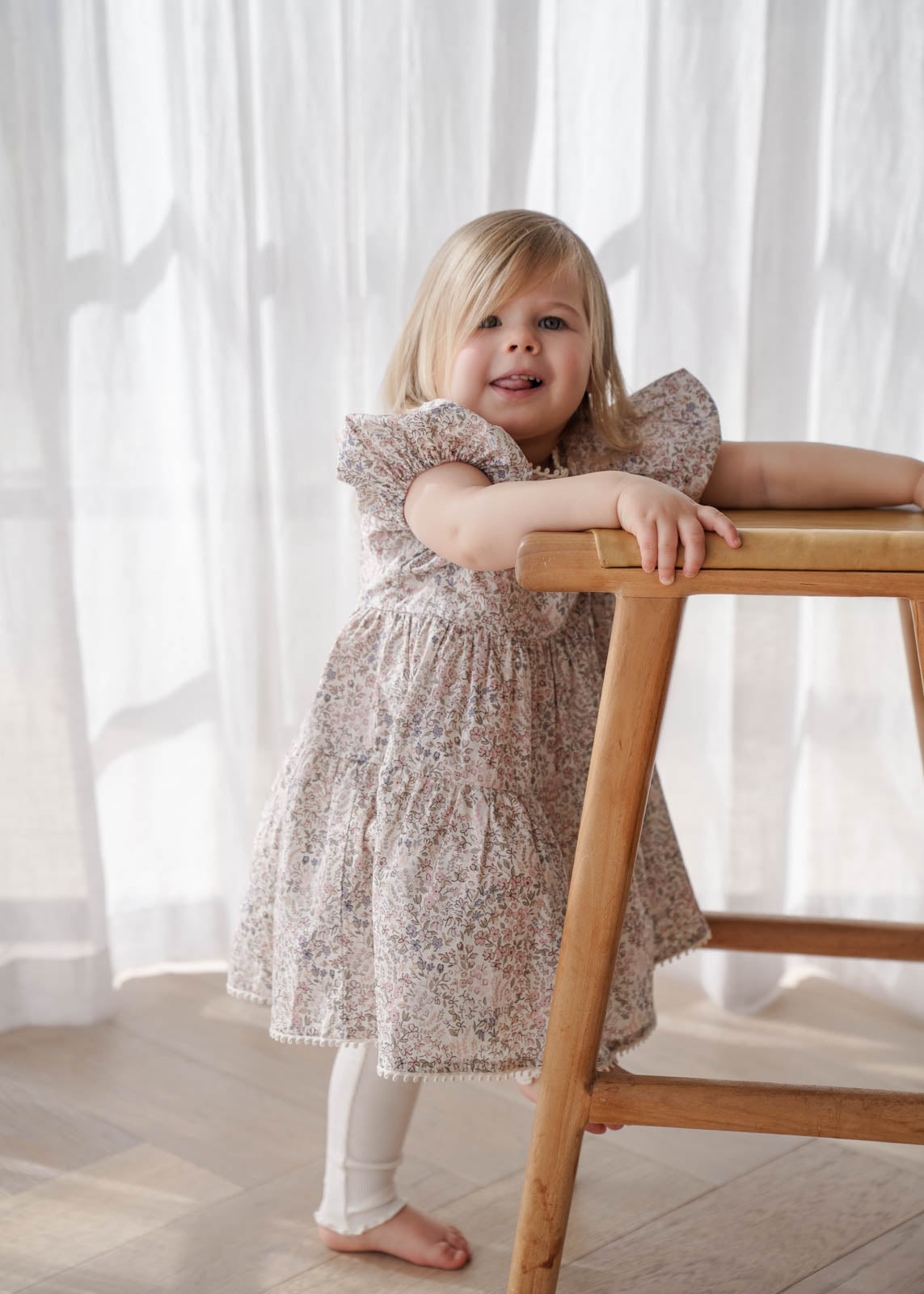 Young girl in a Eliza Floral Dress in Cream standing next to a wooden table with white curtains in the background