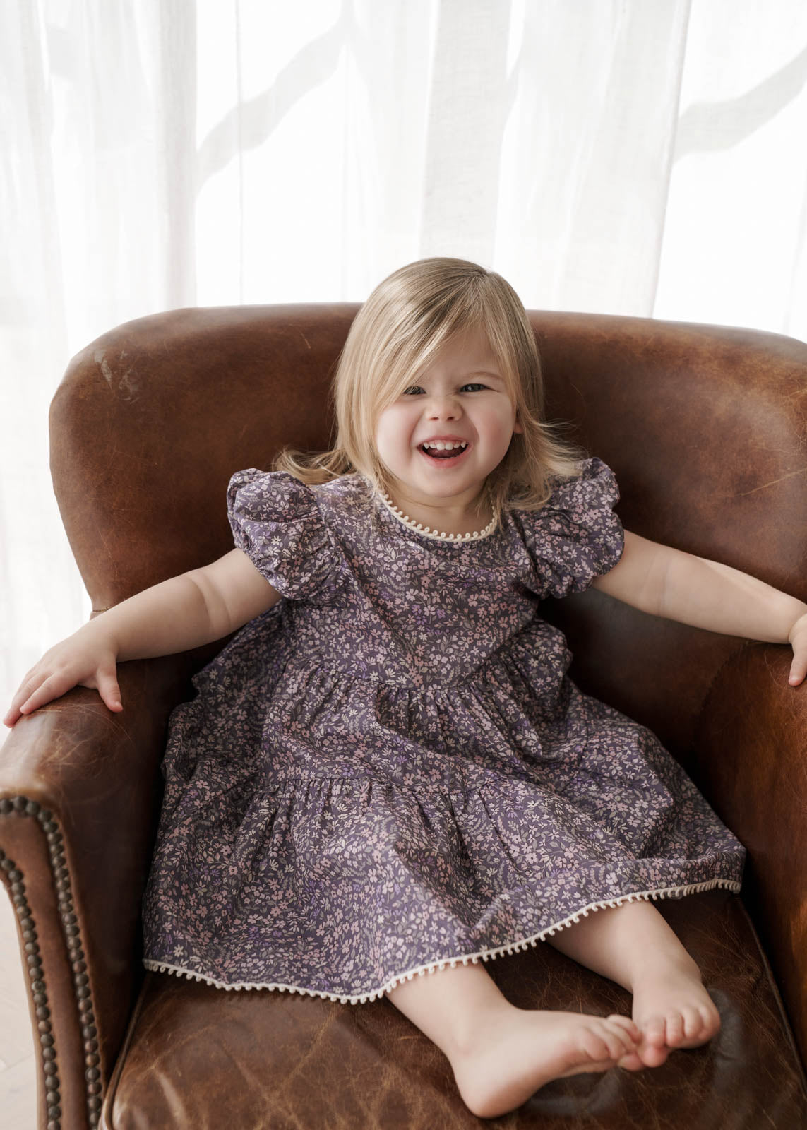 Child in a Eliza Floral Dress in Indigo sitting on a brown armchair with a white curtain background