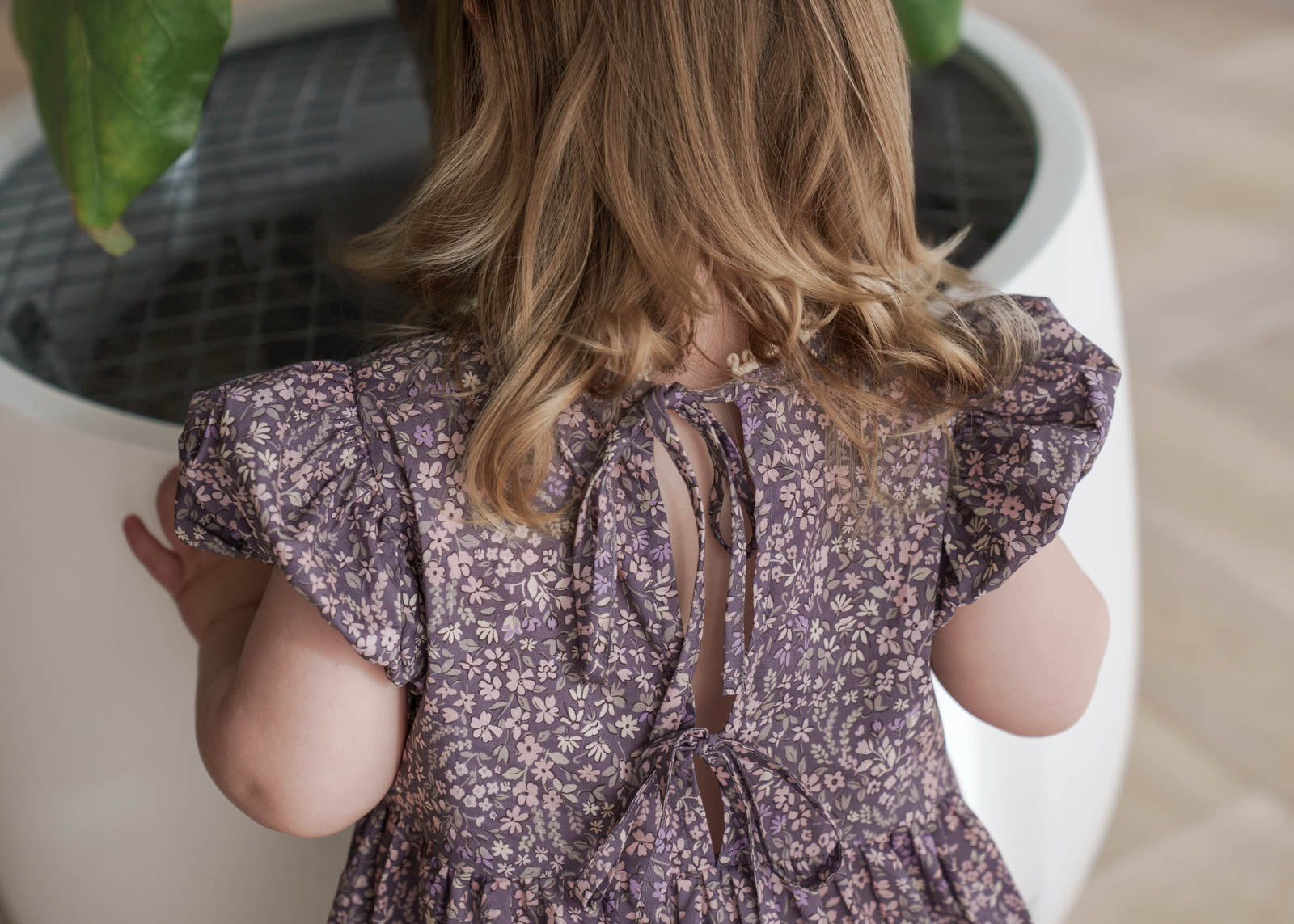 Child wearing a Eliza Floral Dress in Indigo standing next to a white planter with green leaves.