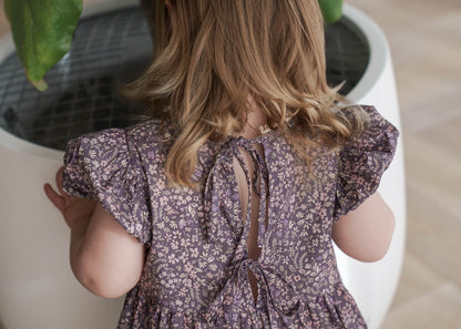 Child wearing a Eliza Floral Dress in Indigo standing next to a white planter with green leaves.