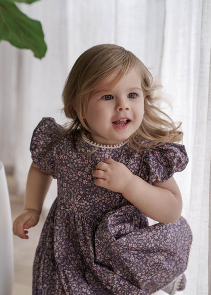 Child wearing a Eliza Floral Dress in Indigo standing indoors with a white curtain background