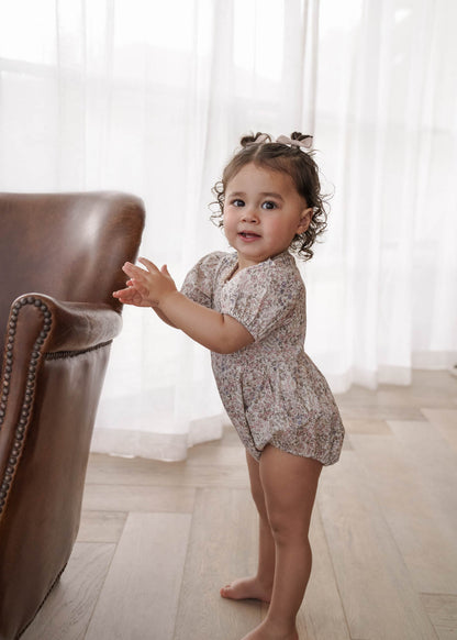 Child in a Eliza Floral Romper in Cream standing near a brown chair with white curtains in the background