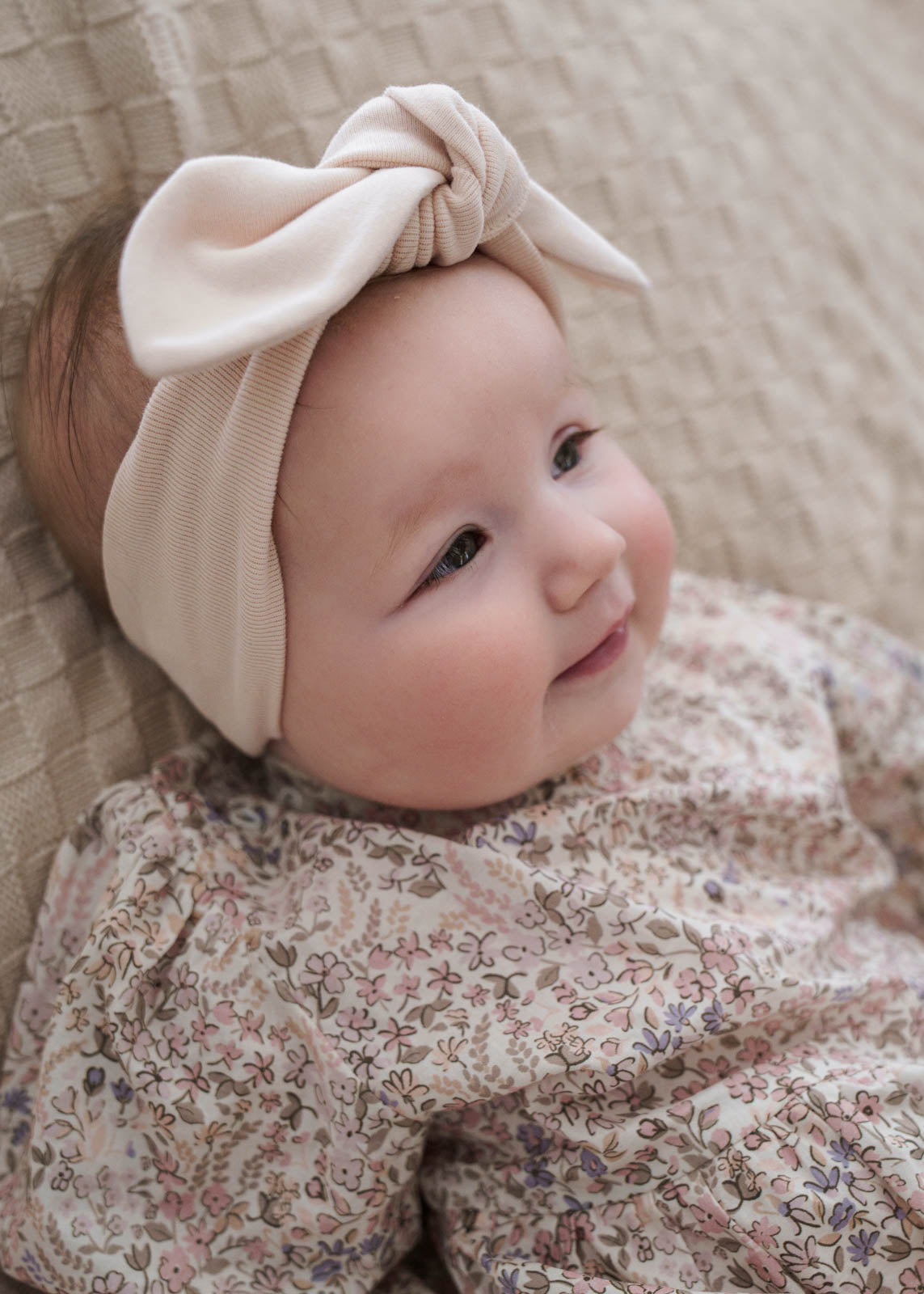 Baby wearing a Eliza Floral Romper in Cream and beige headband with a bow, sitting on a textured surface.