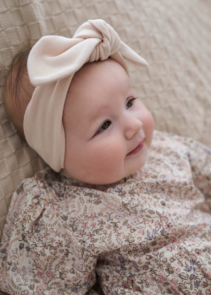 Baby wearing a Eliza Floral Romper in Cream and beige headband with a bow, sitting on a textured surface.