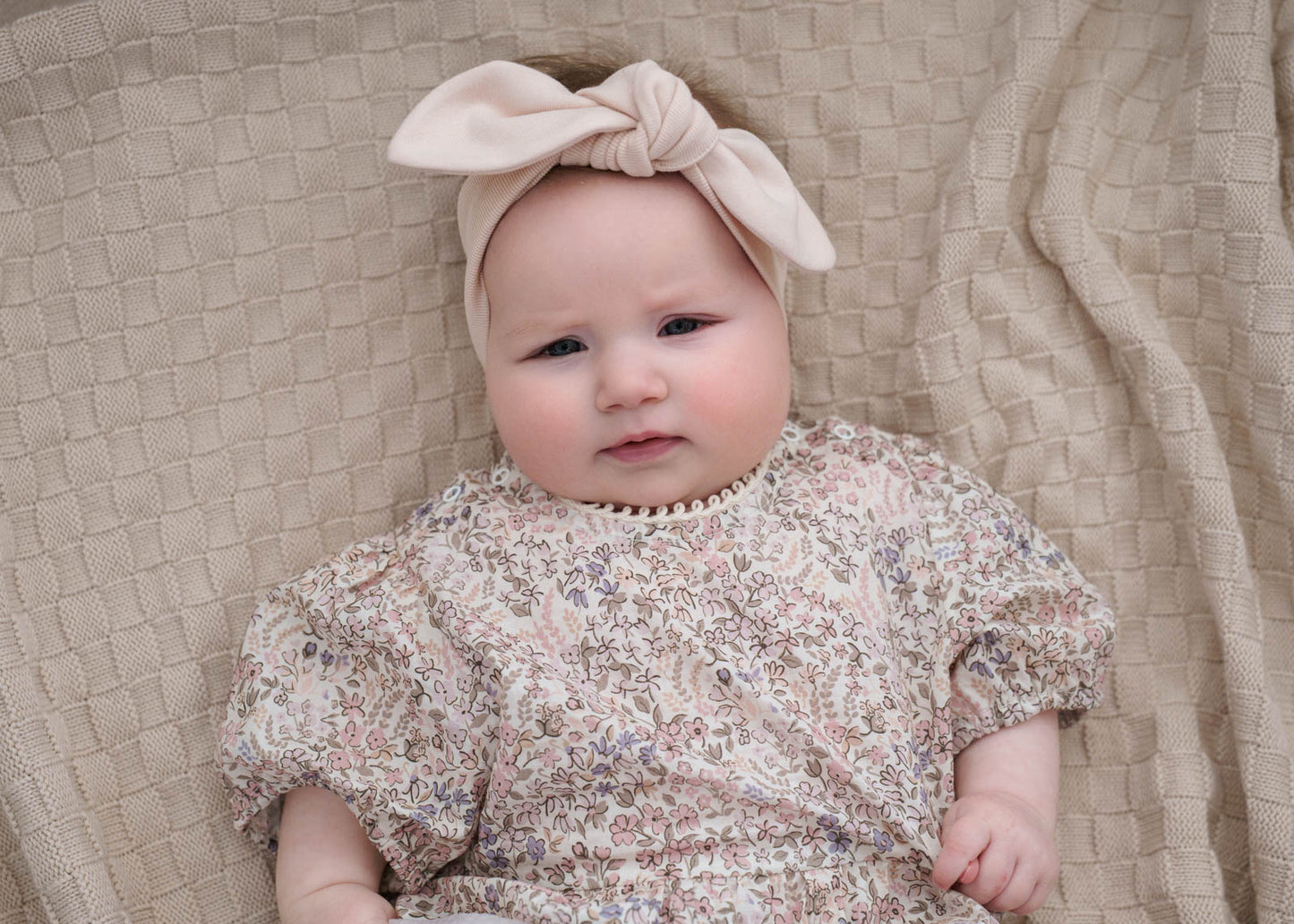 Baby wearing a Eliza Floral Romper in Cream and bow headband on a textured beige blanket