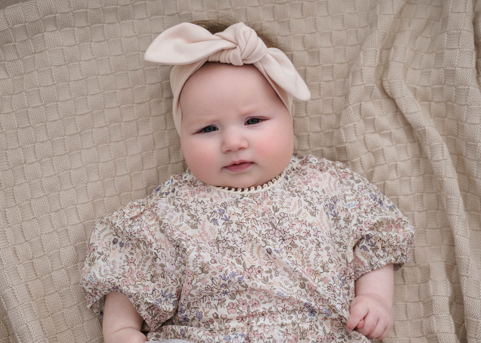 Baby wearing a Eliza Floral Romper in Cream and bow headband on a textured beige blanket