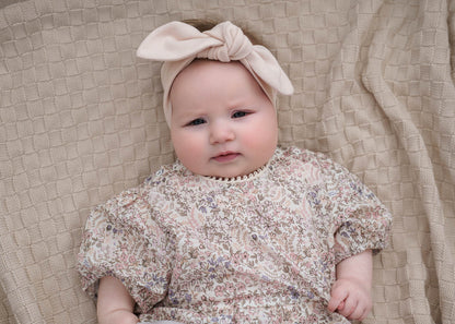 Baby wearing a Eliza Floral Romper in Cream and bow headband on a textured beige blanket