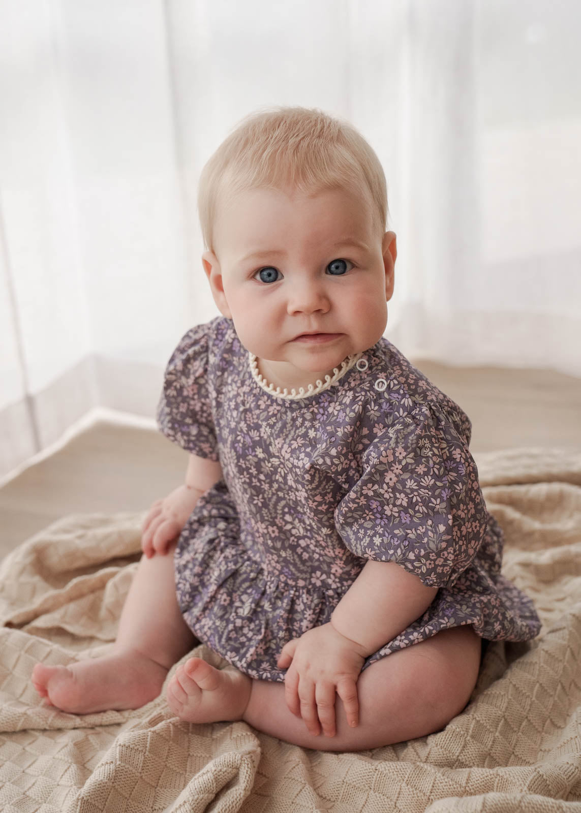 Baby in a Eliza Floral Romper in Indigo sitting on a beige blanket with a white curtain background
