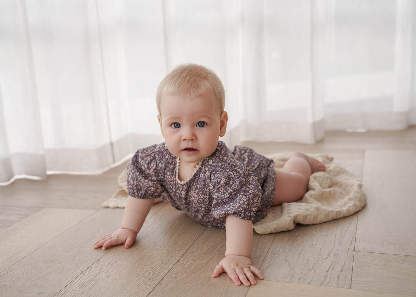 Baby crawling on a wooden floor with a white curtain background, wearing the Eliza Floral Romper in Indigo.