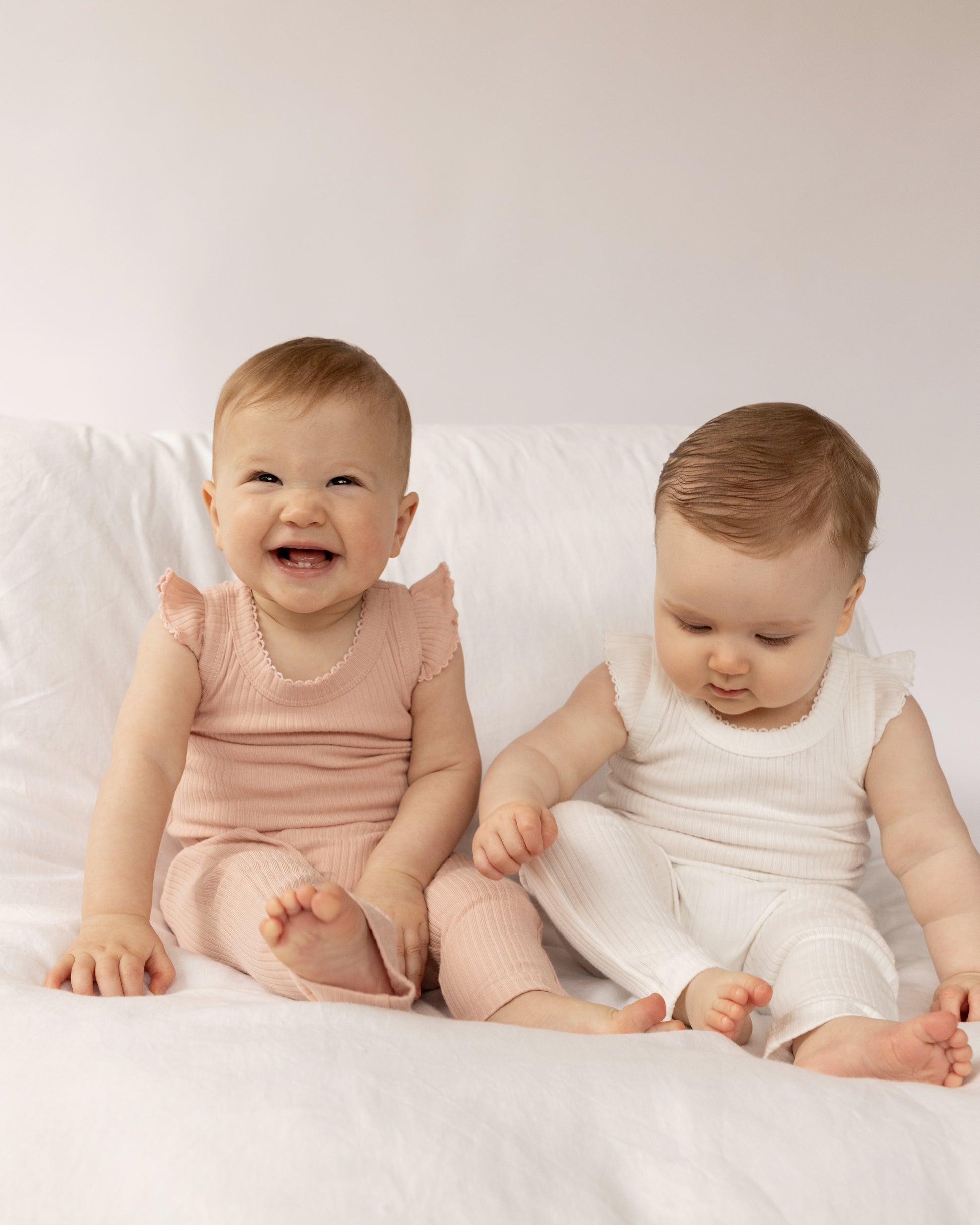 Two babies sitting on a white surface, wearing Frill Basic Singlet Bodysuit in White and Beige on a white bed.