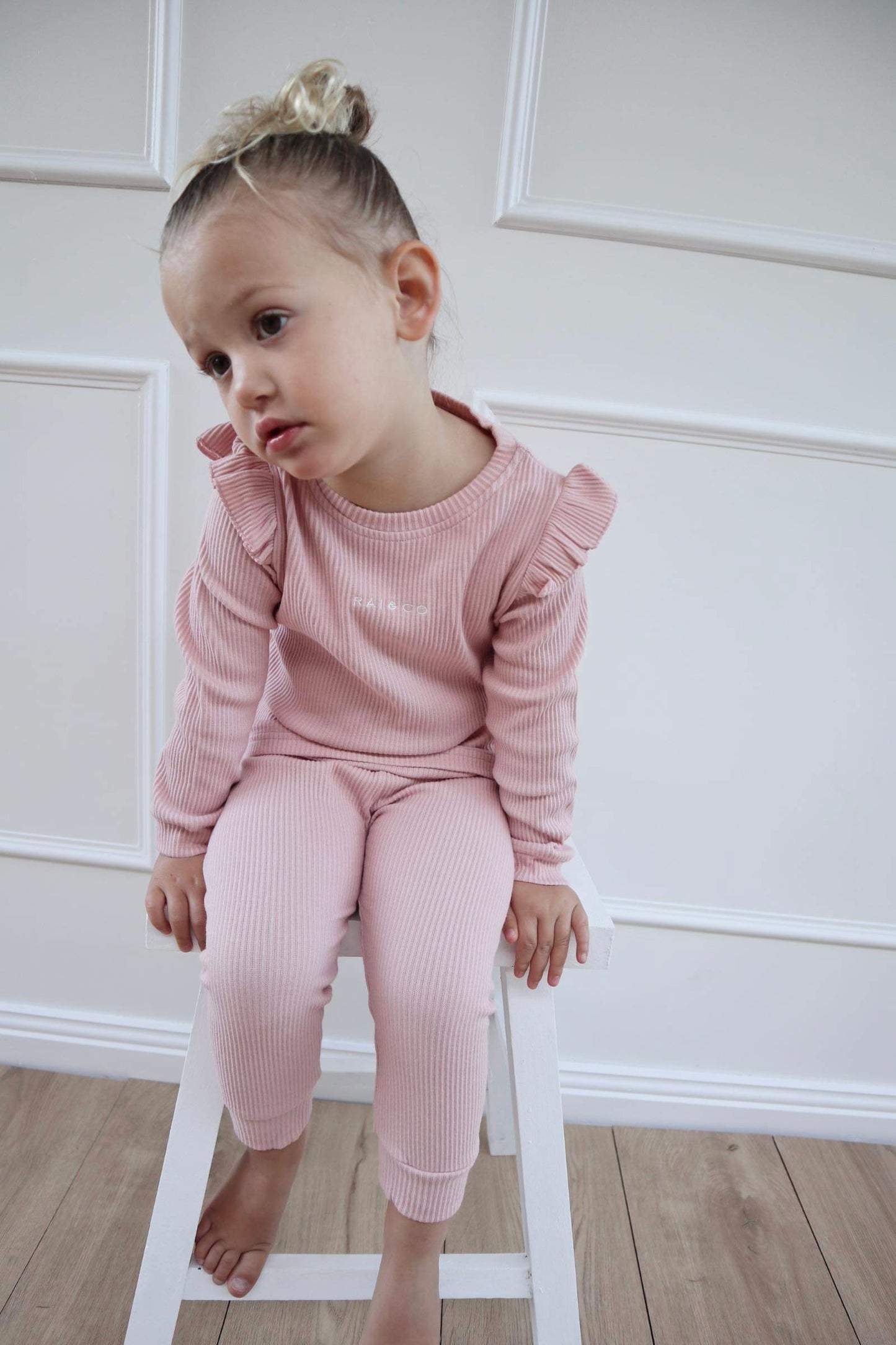 A toddler in wearing Frill Ribbed Two Piece in Powder Pink, sitting against a white background.