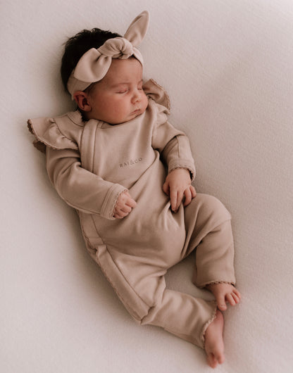 A baby wearing a Frilled Apple Onesie in Dark Beige with a bow on the head, lying on a light background.