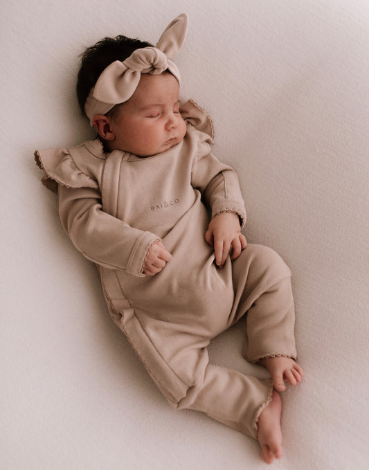 A baby wearing a Frilled Apple Onesie in Dark Beige with a bow on the head, lying on a light background.