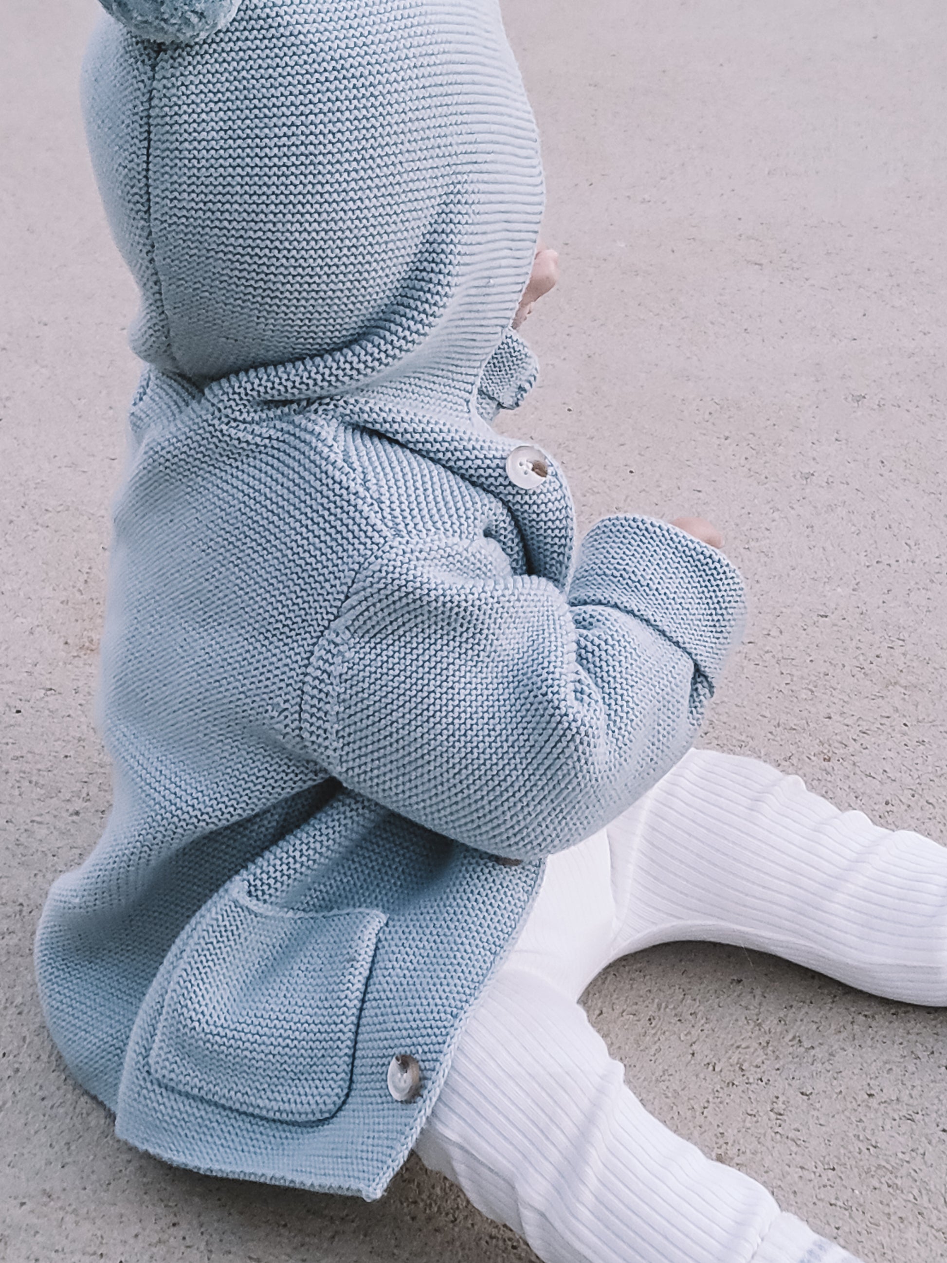 A baby wearing the Hooded Knit in Blue, sitting on the floor.