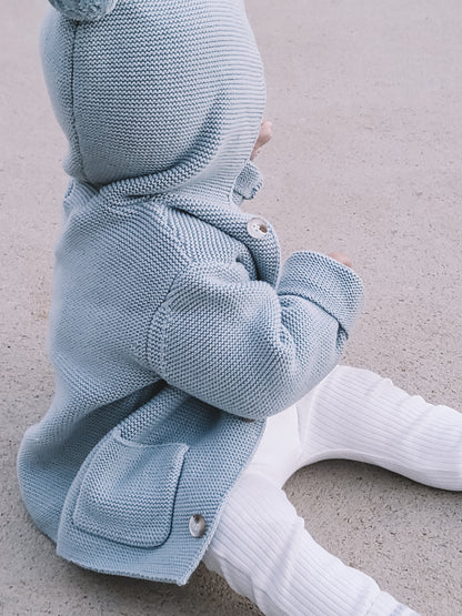 A baby wearing the Hooded Knit in Blue, sitting on the floor.