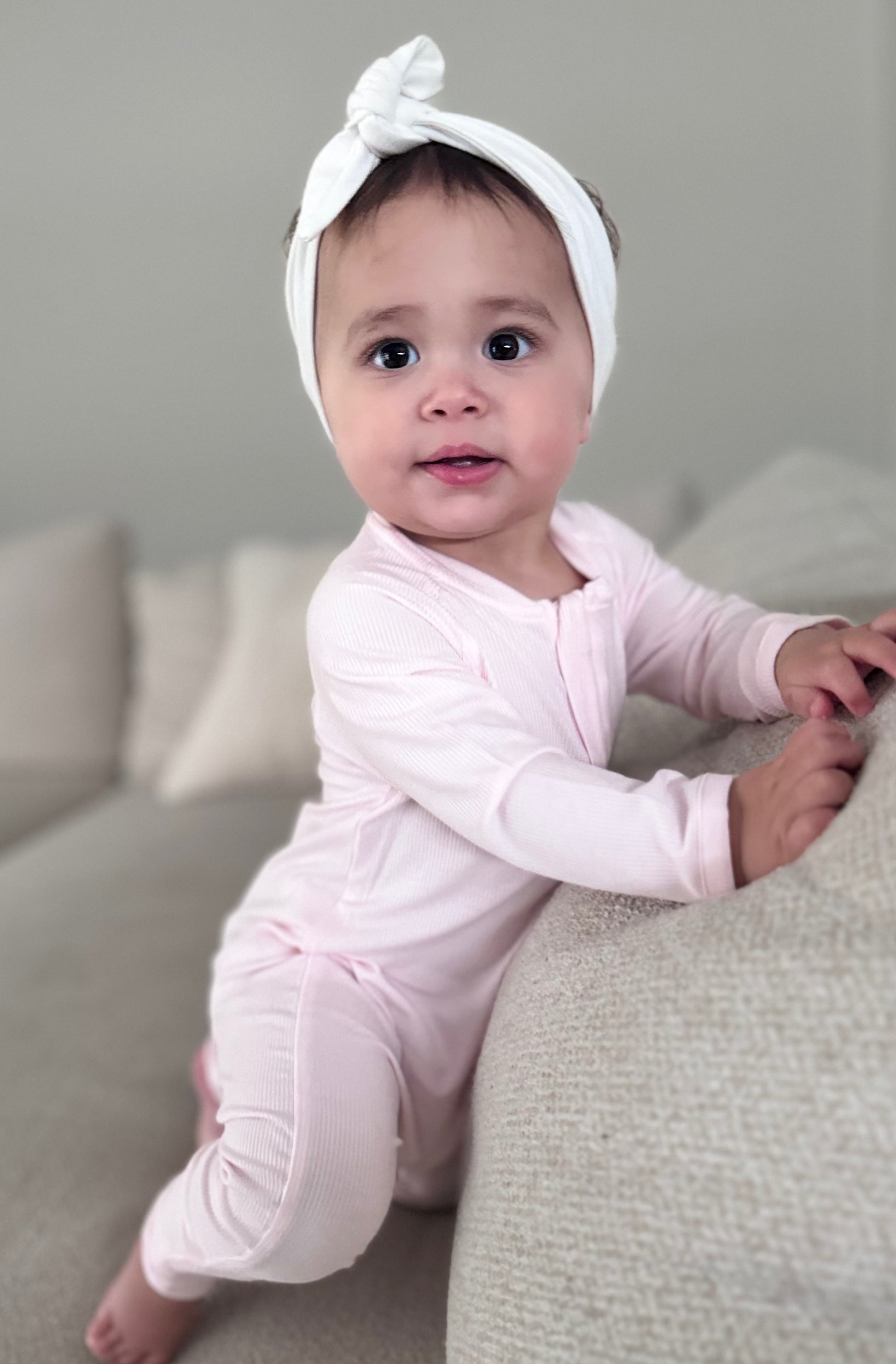 A baby wearing a Lightweight Eco Onesie in Pink with a bow on the head, sitting on a beige couch.