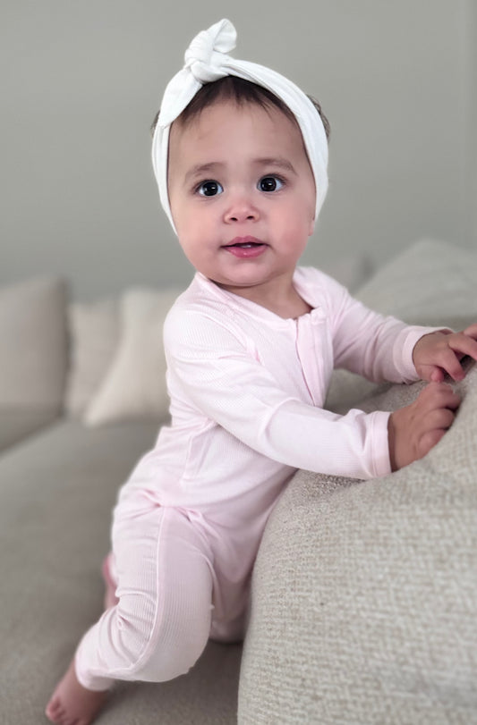 A baby wearing a Lightweight Eco Onesie in Pink with a bow on the head, sitting on a beige couch.