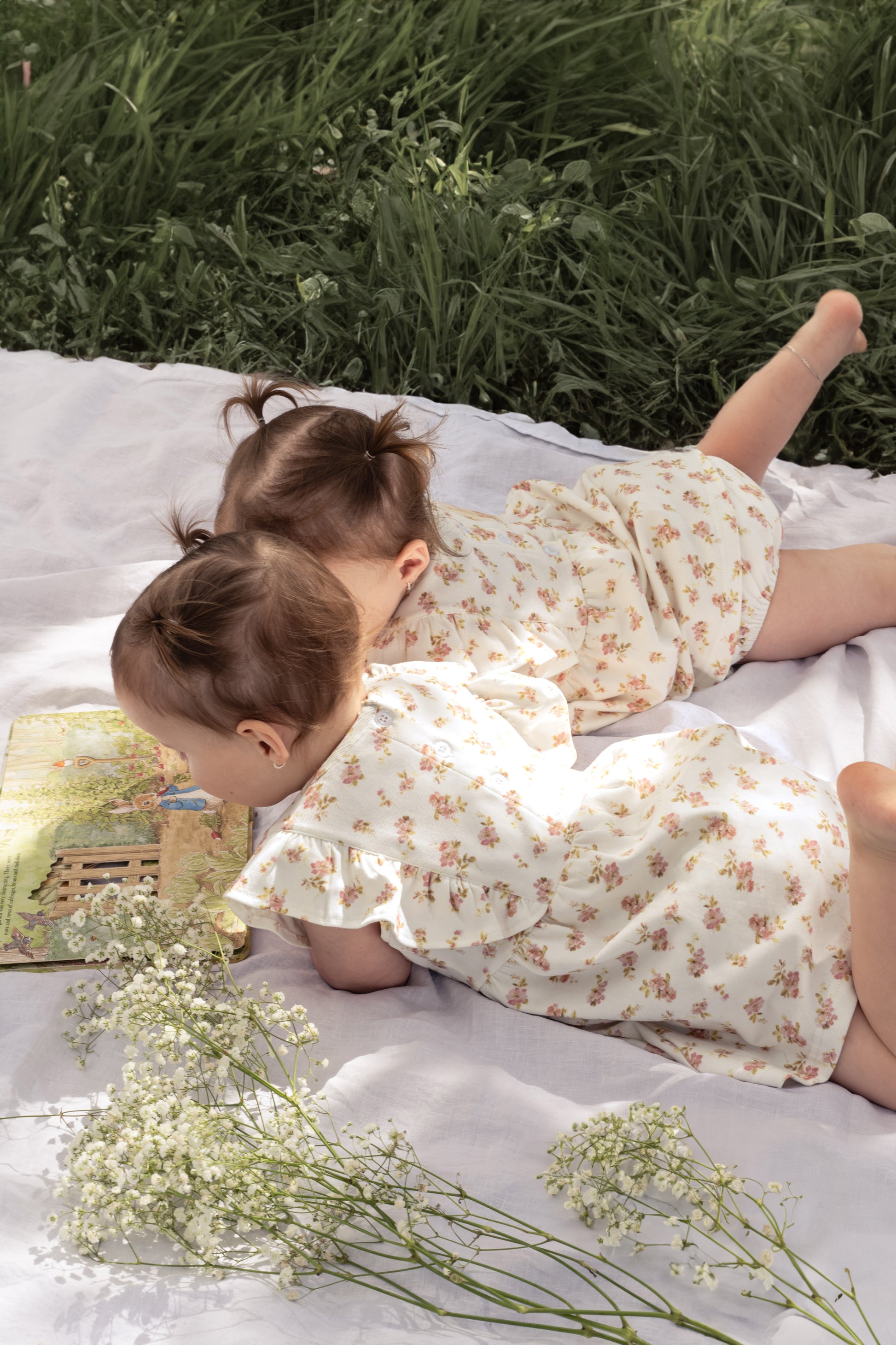 Two young children wearing the Lottie Floral Dress with Pilchers in Pink Floral, lying on a white mat.