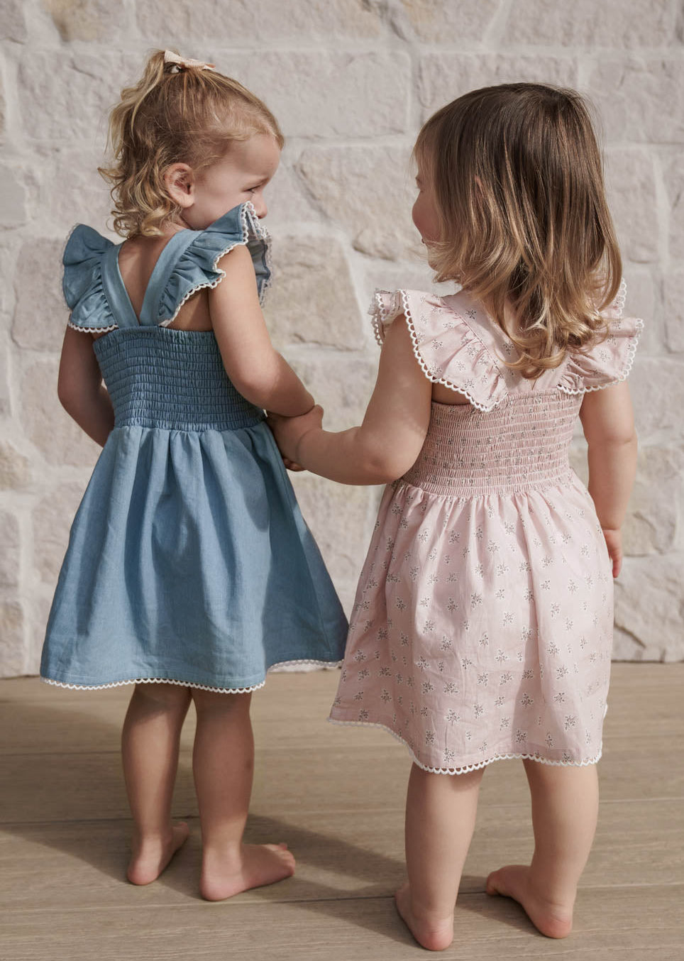 Two young girls in Maddie Dress in Pink Floral and Denim standing close to each other against a stone wall.