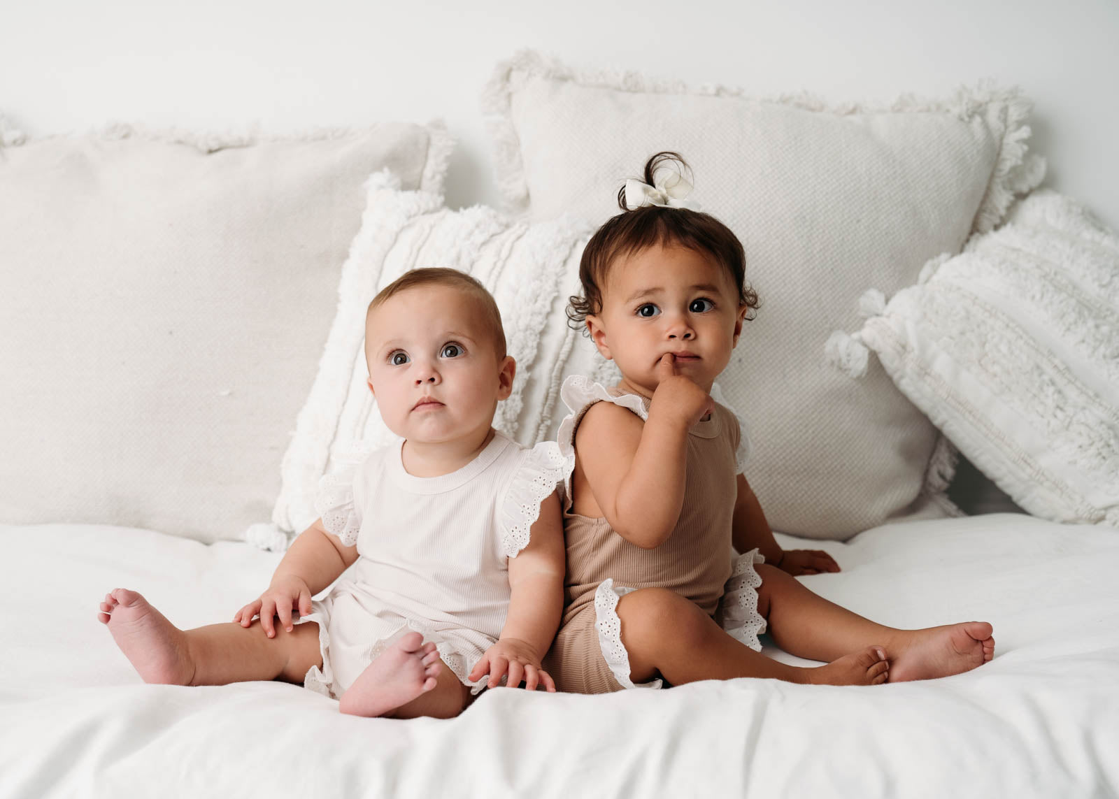 Two babies sitting on a white bed wearing the Mini Ribbed Lace Frill Romper in available colours. 