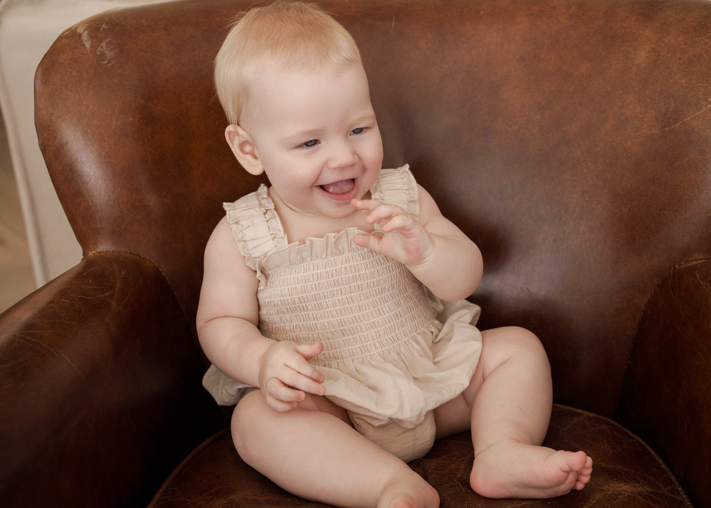 Baby sitting on a brown leather chair, wearing the Ruby Romper in Beige 