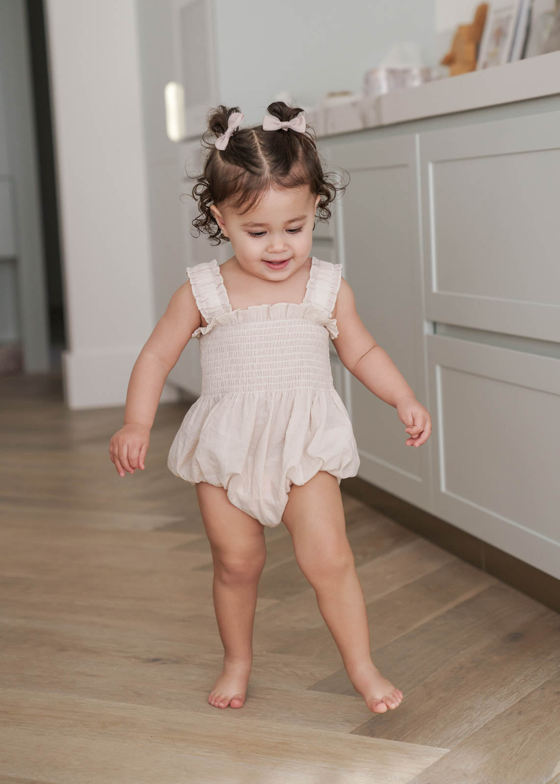 Child in a Ruby Romper in Beige standing on a wooden floor in a kitchen. 