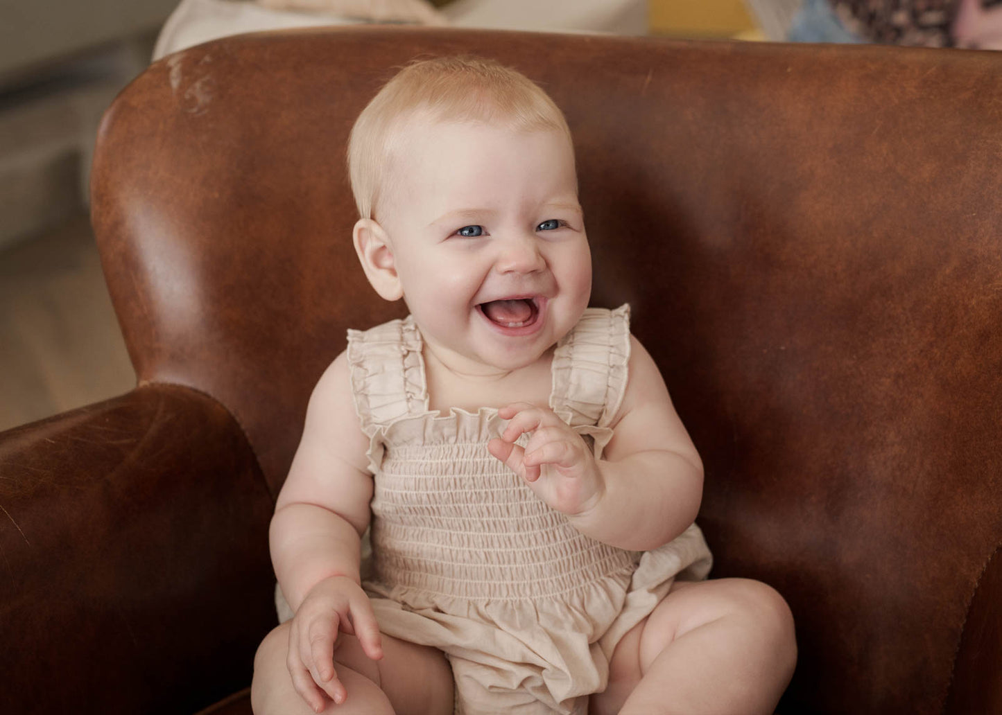 Baby sitting on a brown leather chair with a happy expression wearing Ruby Romper in Beige