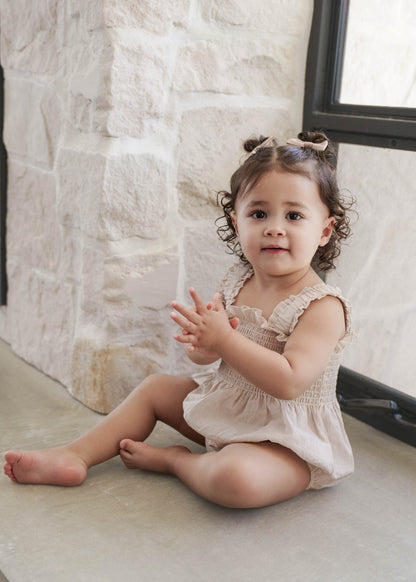 Young child sitting on a stone floor against a neutral background, wearing the Ruby Romper in Beige 