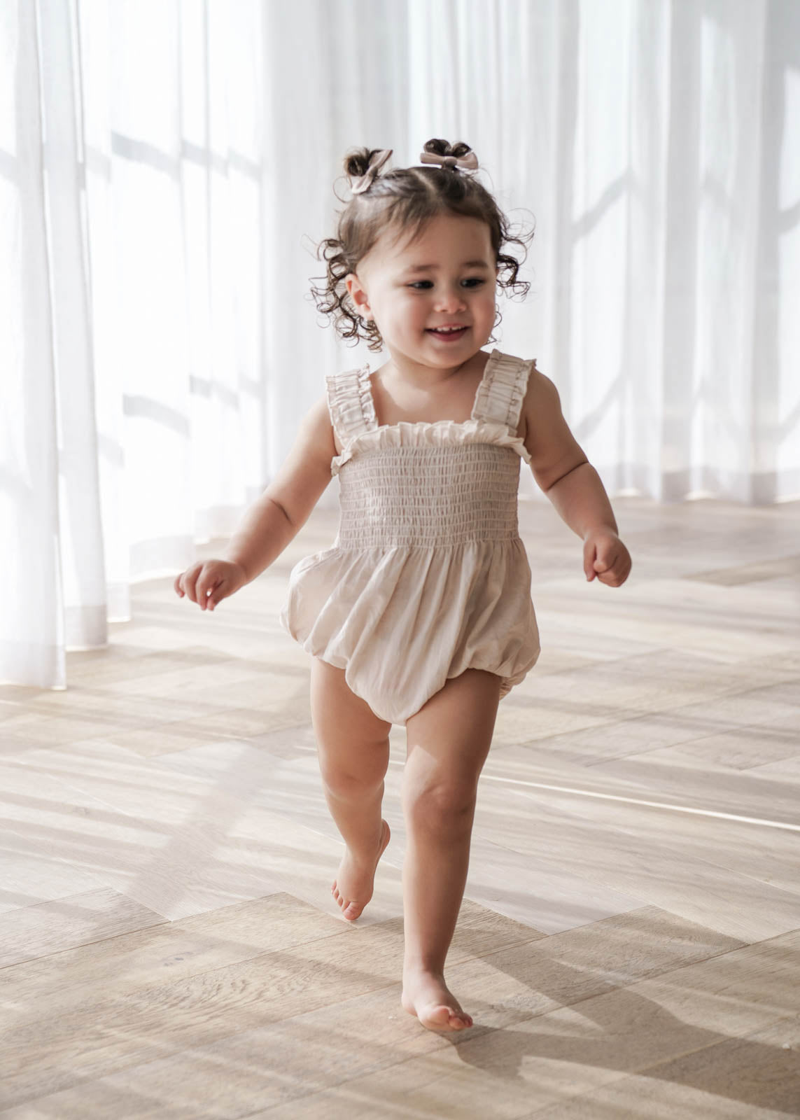 Child in a Beige Ruby Romper walking on a wooden floor with white curtains in the background
