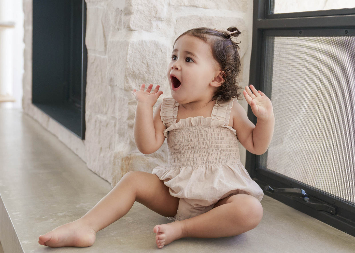 Baby in a Beige Ruby Romper sitting on a floor with a neutral background