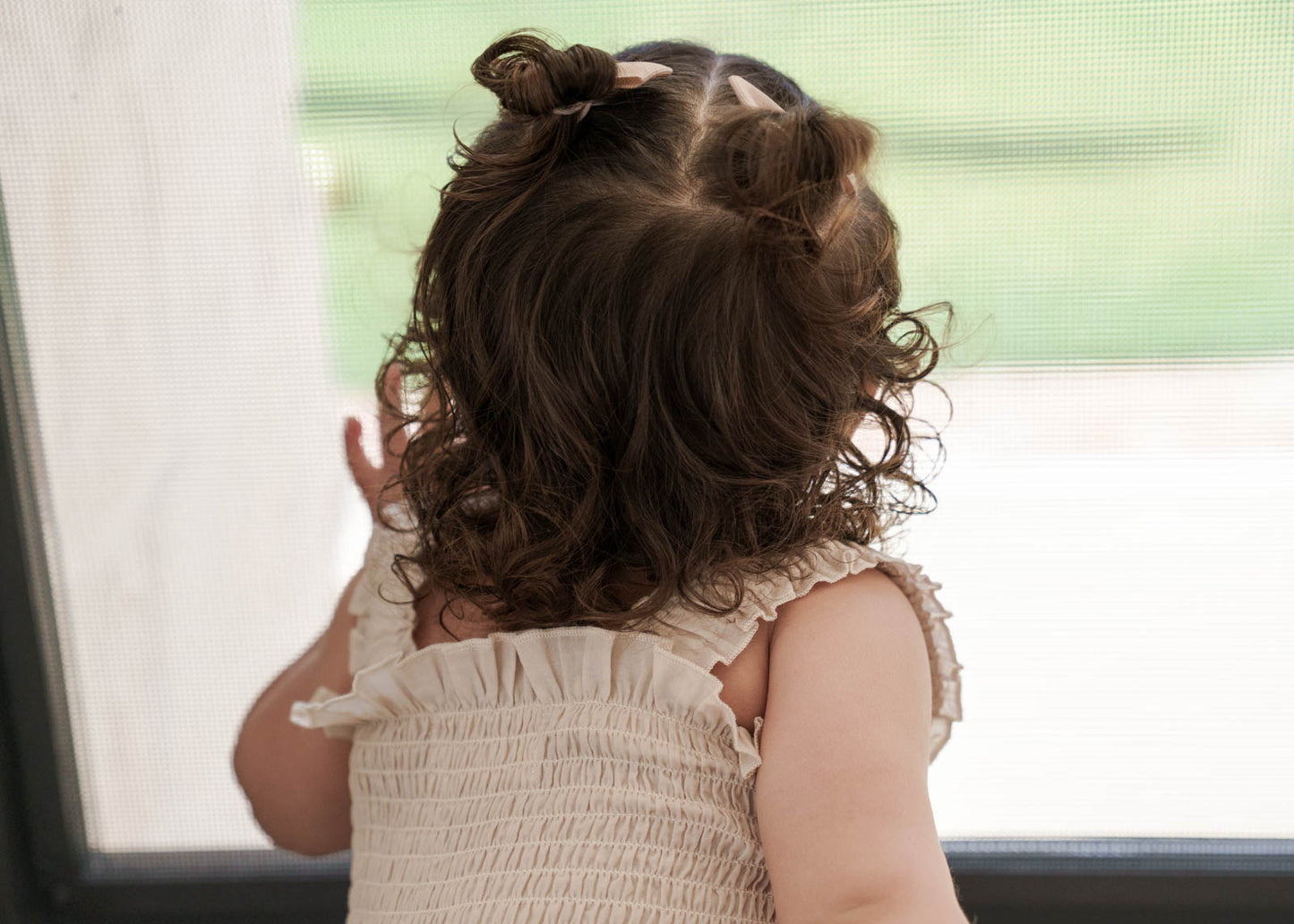 Child with styled hair looking out of a window, wearing the Ruby Romper in Beige