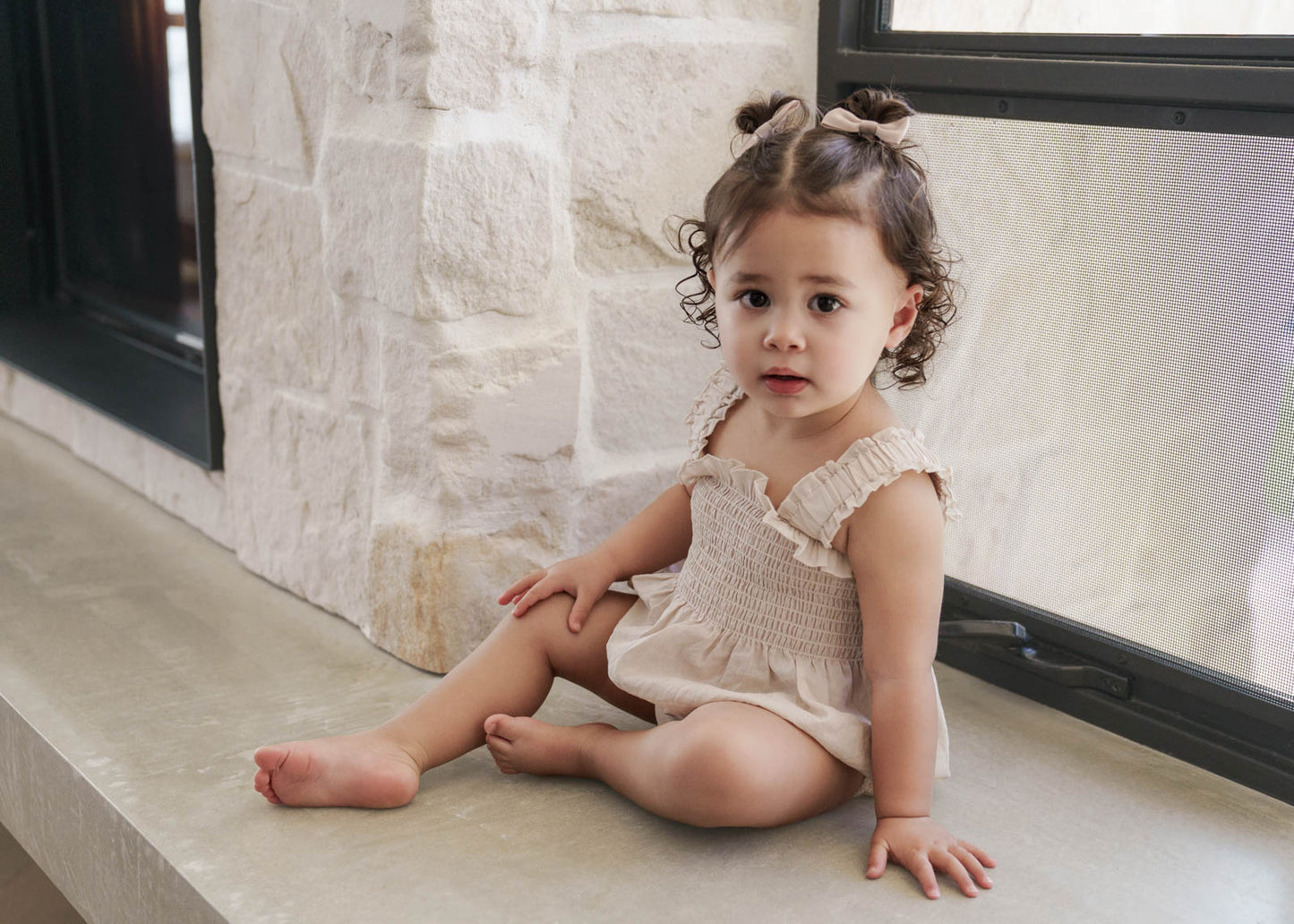 Young child sitting on a windowsill with a stone wall background, wearing the Ruby Romper in Beige 