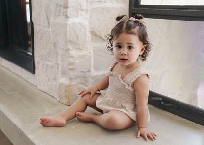 Young child sitting on a windowsill with a stone wall background, wearing the Ruby Romper in Beige 