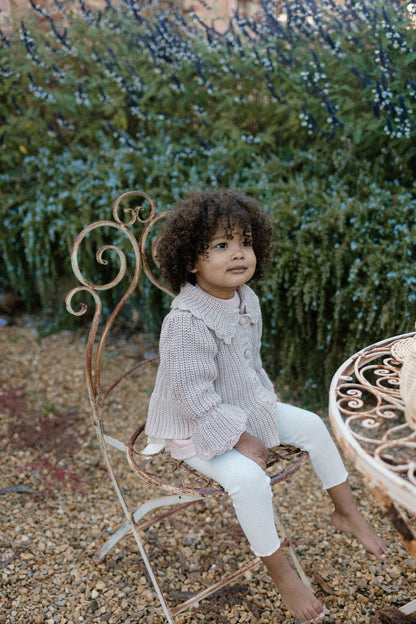 A young girl sitting on a chair outdoors wearing a Kenya Cardigan