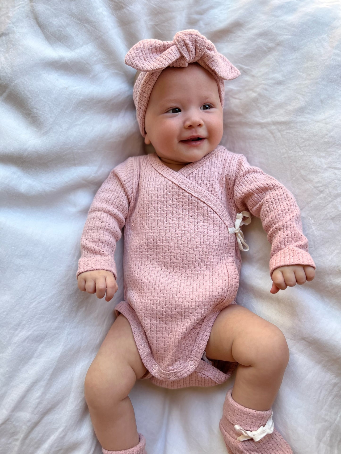 Baby wearing a Knitted Knot Headband in Pink on a white bed