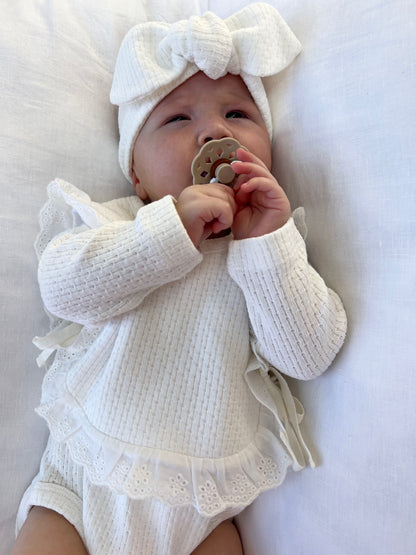 A baby wearing a Knitted Knot Headband in White, lying down and chewing on a pacifier.