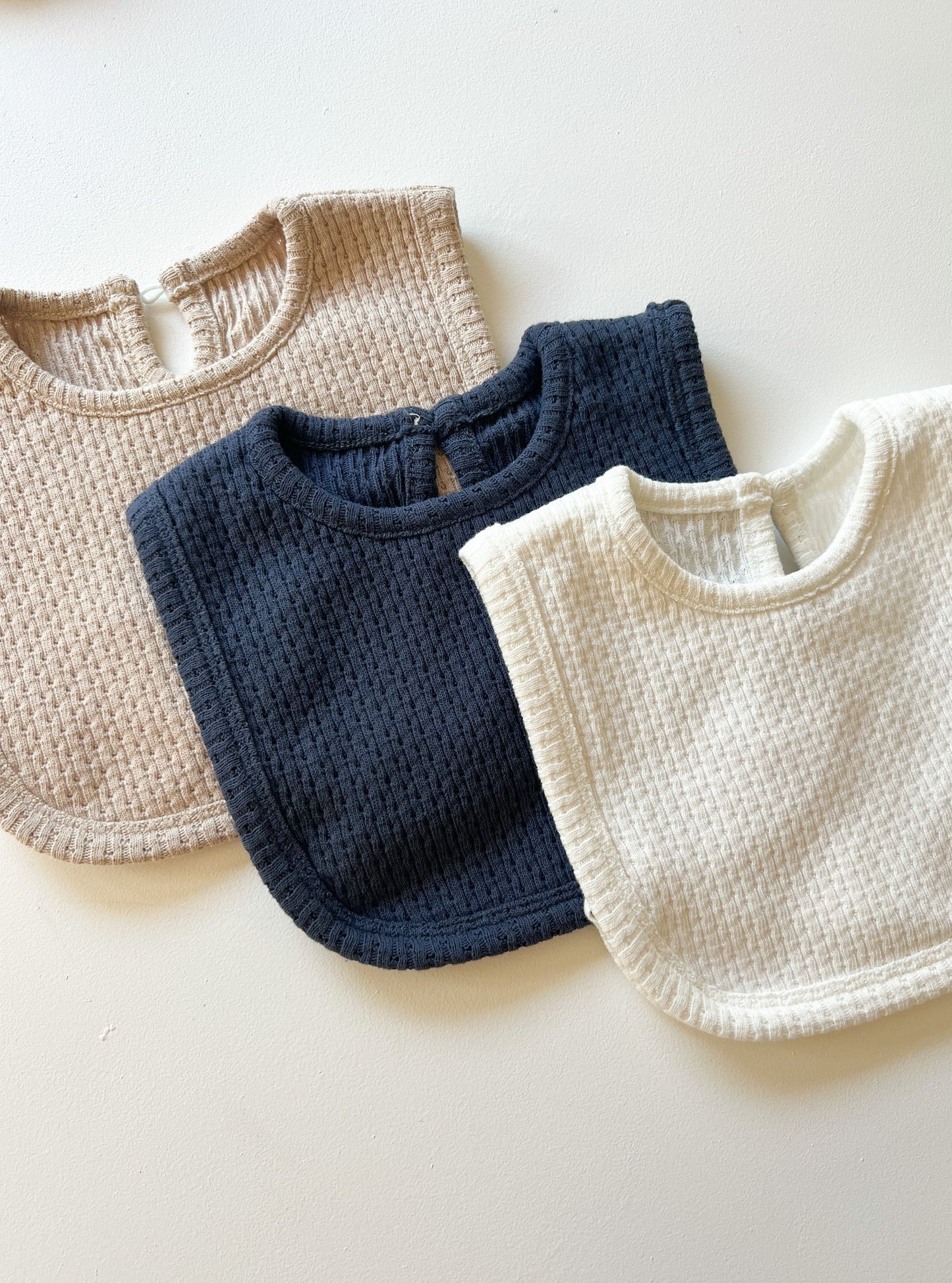 Three Knitted Plain Bib in Available Colours laid out flat in a horizontal arrangement. 