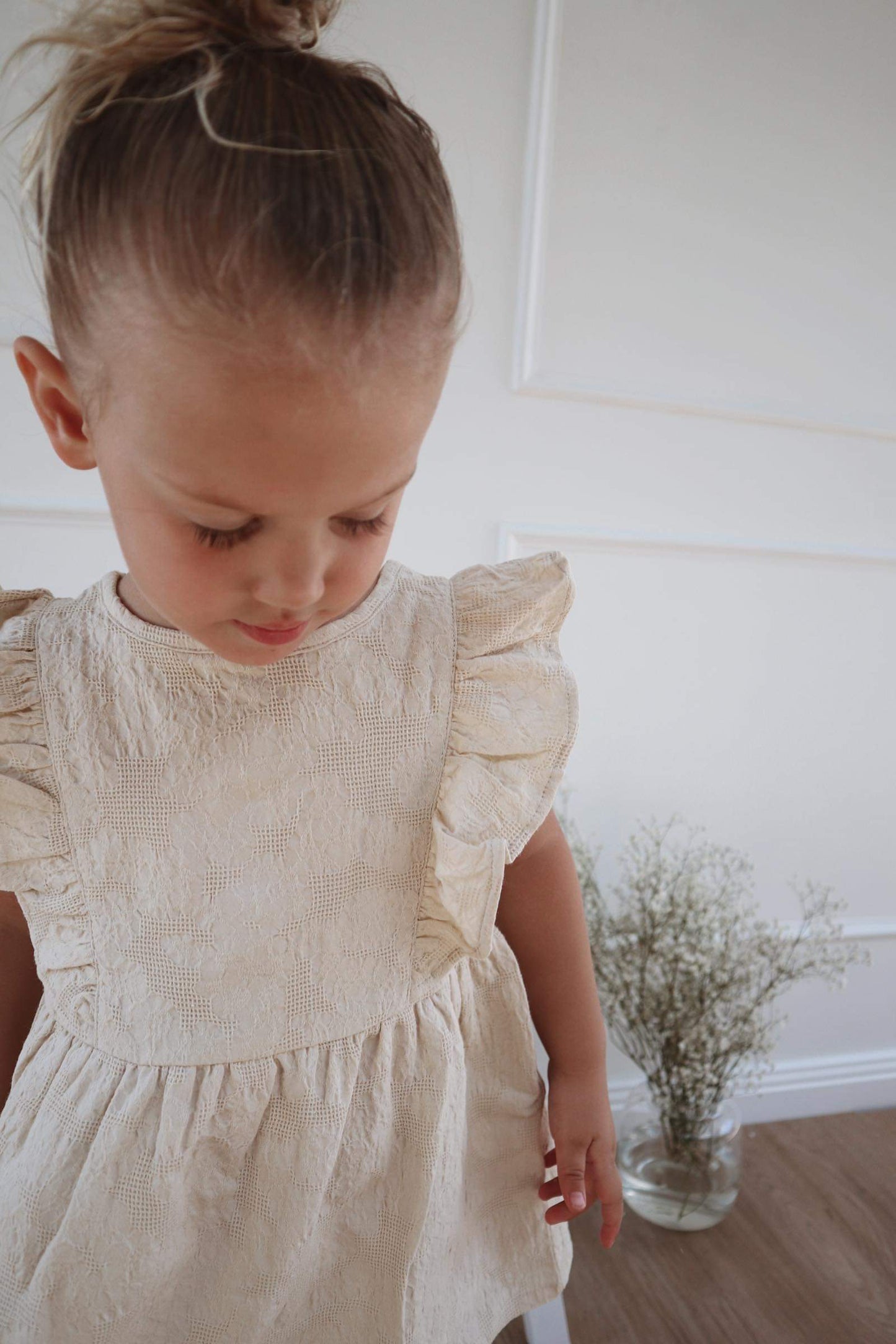 A young child wearing a Lou Lou Frilled Dress With Pilchers in Beige, standing indoors.