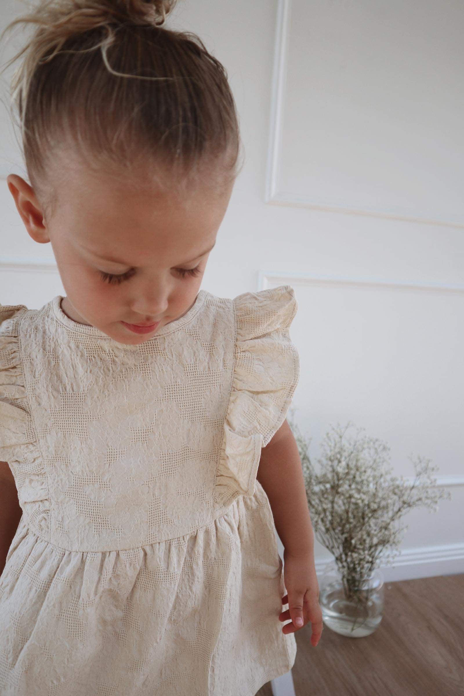 A young child wearing a Lou Lou Frilled Dress With Pilchers in Beige, standing indoors.